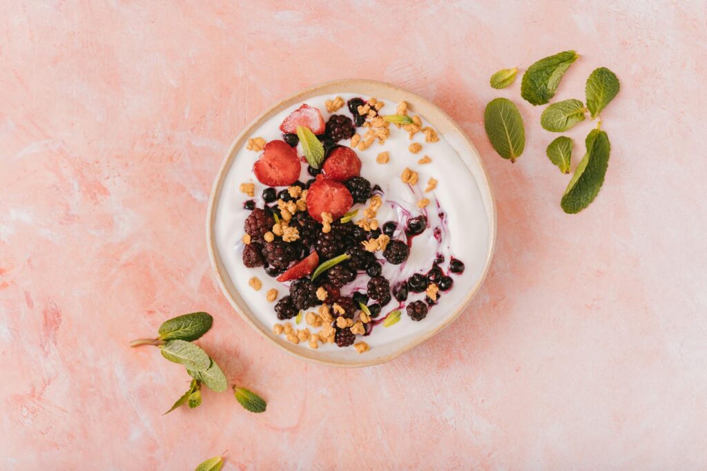 Top view of a vibrant yogurt bowl with mixed berries, granola, and mint leaves on a pink background.